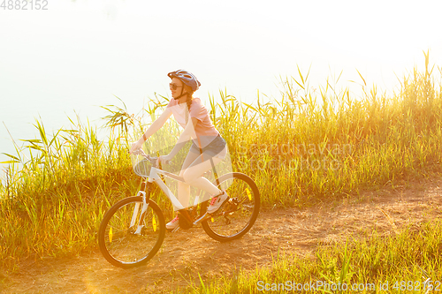 Image of Joyful young woman riding a bicycle at the riverside and meadow promenade