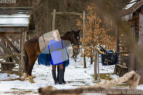 Image of A beautiful girl in a blue dress stands with a horse against the background of old wooden buildings in a winter forest