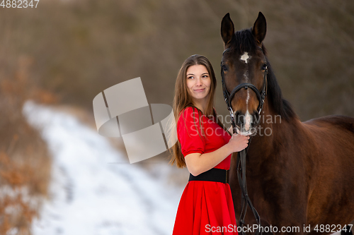 Image of Portrait of a beautiful girl in a red dress standing with a horse on the background of a winter road