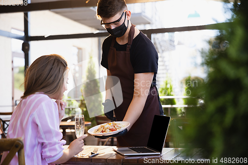 Image of The waiter works in a restaurant in a medical mask, gloves during coronavirus pandemic