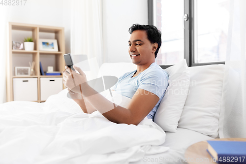 Image of happy indian man with smartphone in bed at home