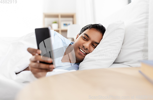 Image of happy indian man with smartphone in bed at home