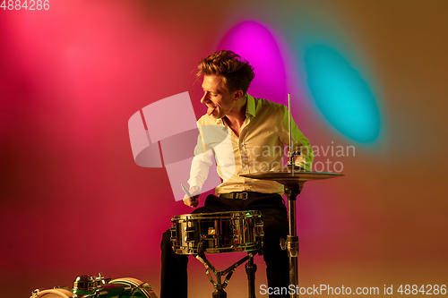 Image of Young caucasian musician, drummer playing on gradient background in neon light. Concept of music, hobby, festival