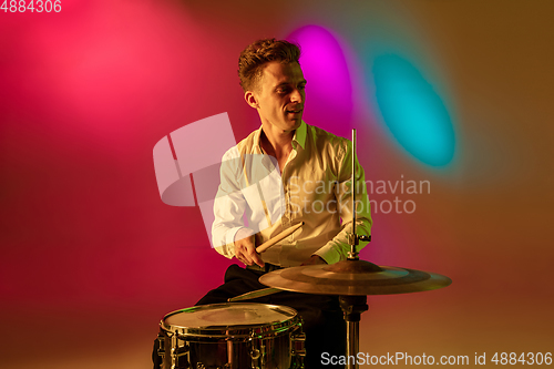 Image of Young caucasian musician, drummer playing on gradient background in neon light. Concept of music, hobby, festival