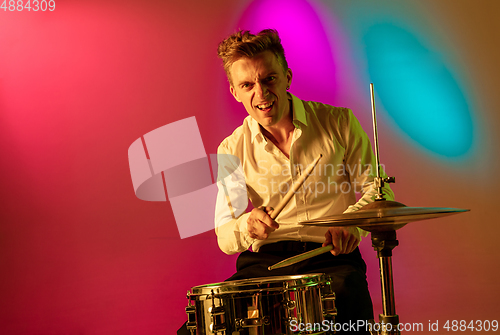 Image of Young caucasian musician, drummer playing on gradient background in neon light. Concept of music, hobby, festival