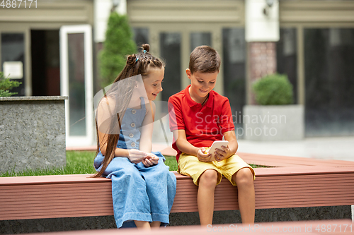 Image of Two smiling kids, boy and girl playing smartphone together in town, city in summer day