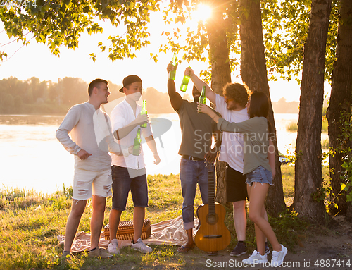 Image of Group of friends clinking beer bottles during picnic at the beach. Lifestyle, friendship, having fun, weekend and resting concept.