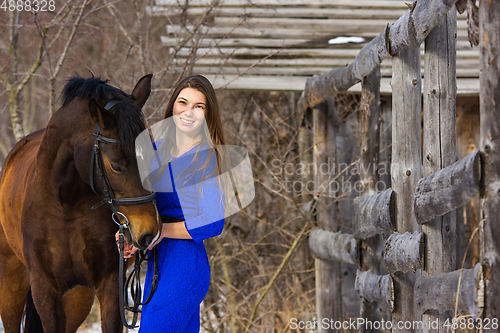 Image of A beautiful young girl in a blue dress hugs a horse against the background of an old fence and a winter forest