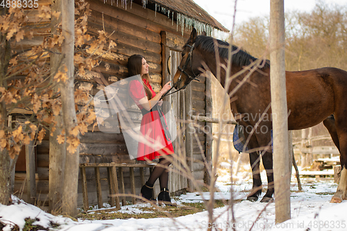 Image of A girl in a red dress stands near an old wooden house, a horse stands next to her.