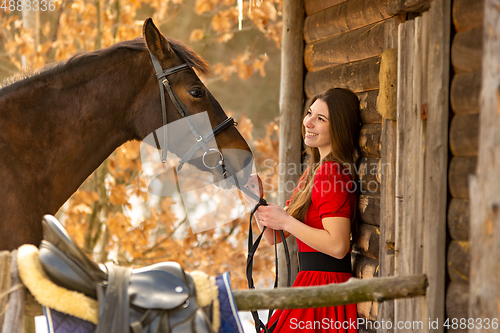 Image of Portrait of a beautiful girl in a red dress, a girl holding a horse by the bridle, a beautiful background of a wooden wall and forest