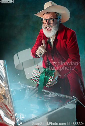 Image of Modern stylish Santa Claus in red fashionable suit and cowboy\'s hat on dark background