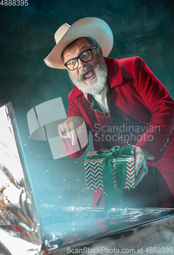 Image of Modern stylish Santa Claus in red fashionable suit and cowboy\'s hat on dark background