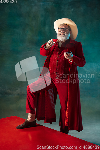 Image of Modern stylish Santa Claus in red fashionable suit and cowboy\'s hat on dark background