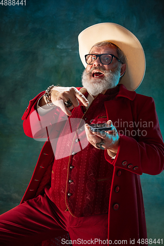 Image of Modern stylish Santa Claus in red fashionable suit and cowboy\'s hat on dark background