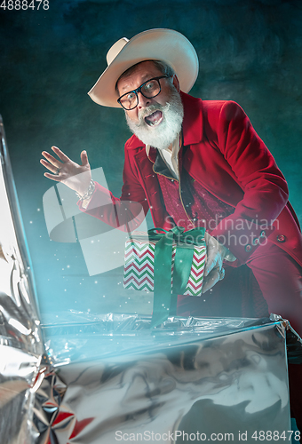 Image of Modern stylish Santa Claus in red fashionable suit and cowboy\'s hat on dark background