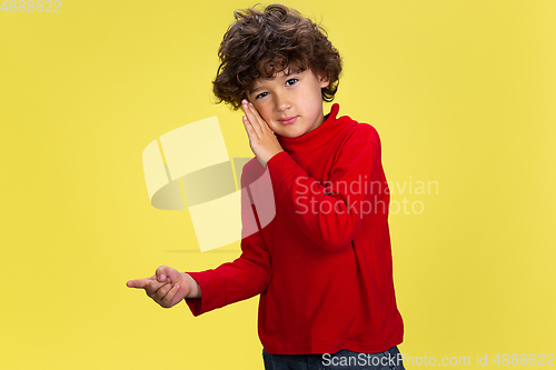 Image of Pretty young curly boy in red wear on yellow studio background. Childhood, expression, fun.