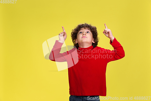 Image of Pretty young curly boy in red wear on yellow studio background. Childhood, expression, fun.