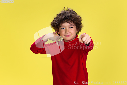 Image of Pretty young curly boy in red wear on yellow studio background. Childhood, expression, fun.