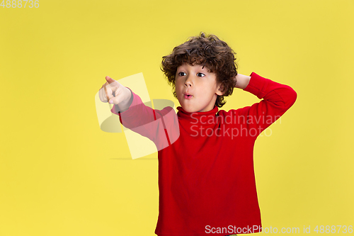 Image of Pretty young curly boy in red wear on yellow studio background. Childhood, expression, fun.