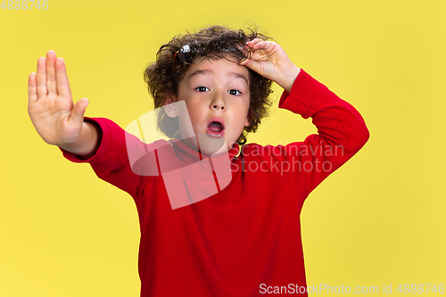 Image of Pretty young curly boy in red wear on yellow studio background. Childhood, expression, fun.