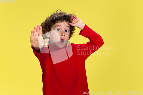 Image of Pretty young curly boy in red wear on yellow studio background. Childhood, expression, fun.