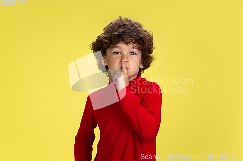 Image of Pretty young curly boy in red wear on yellow studio background. Childhood, expression, fun.