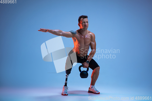 Image of Athlete with disabilities or amputee isolated on blue studio background. Professional male sportsman with leg prosthesis training with weights in neon