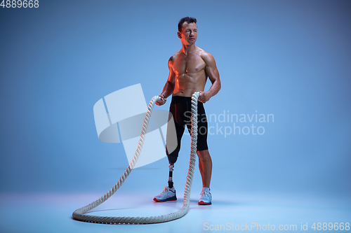 Image of Athlete with disabilities or amputee isolated on blue studio background. Professional male sportsman with leg prosthesis training with weights in neon