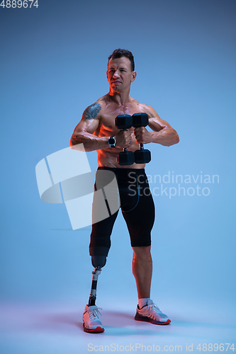 Image of Athlete with disabilities or amputee isolated on blue studio background. Professional male sportsman with leg prosthesis training with weights in neon