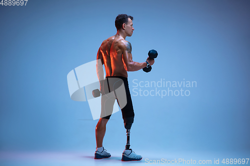 Image of Athlete with disabilities or amputee isolated on blue studio background. Professional male sportsman with leg prosthesis training with weights in neon