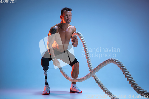 Image of Athlete with disabilities or amputee isolated on blue studio background. Professional male sportsman with leg prosthesis training with weights in neon