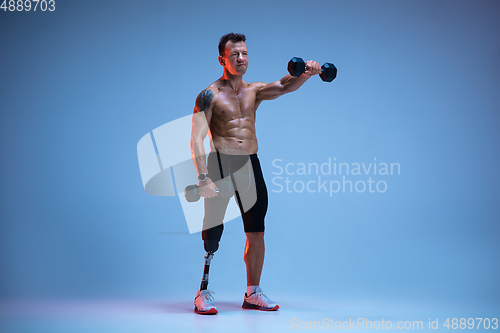 Image of Athlete with disabilities or amputee isolated on blue studio background. Professional male sportsman with leg prosthesis training with weights in neon