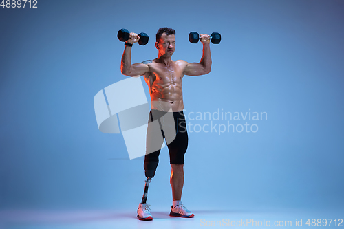 Image of Athlete with disabilities or amputee isolated on blue studio background. Professional male sportsman with leg prosthesis training with weights in neon