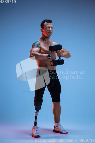 Image of Athlete with disabilities or amputee isolated on blue studio background. Professional male sportsman with leg prosthesis training with weights in neon