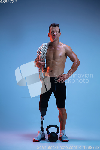 Image of Athlete with disabilities or amputee isolated on blue studio background. Professional male sportsman with leg prosthesis training with weights in neon