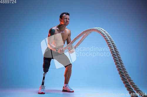 Image of Athlete with disabilities or amputee isolated on blue studio background. Professional male sportsman with leg prosthesis training with weights in neon