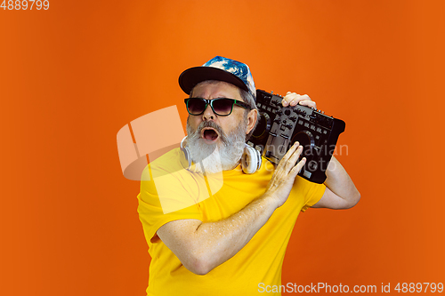 Image of Senior hipster man using devices, gadgets on orange background. Tech and joyful elderly lifestyle concept