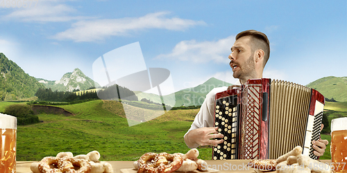 Image of The happy smiling man with beer dressed in traditional Austrian or Bavarian costume holding mug of beer, mountains on background, flyer