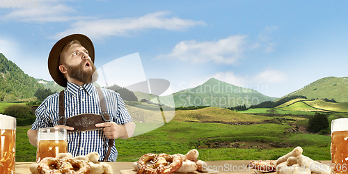 Image of The happy smiling man with beer dressed in traditional Austrian or Bavarian costume holding mug of beer, mountains on background, flyer