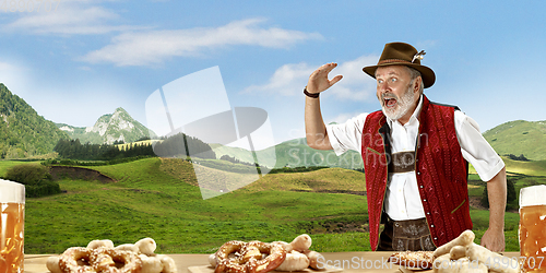 Image of The senior happy smiling man with beer dressed in traditional Austrian or Bavarian costume holding mug of beer, mountains on background, flyer