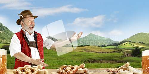 Image of The senior happy smiling man with beer dressed in traditional Austrian or Bavarian costume holding mug of beer, mountains on background, flyer