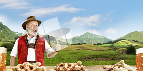 Image of The senior happy smiling man with beer dressed in traditional Austrian or Bavarian costume holding mug of beer, mountains on background, flyer