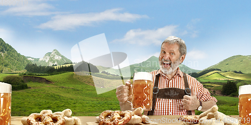 Image of The senior happy smiling man with beer dressed in traditional Austrian or Bavarian costume holding mug of beer, mountains on background, flyer