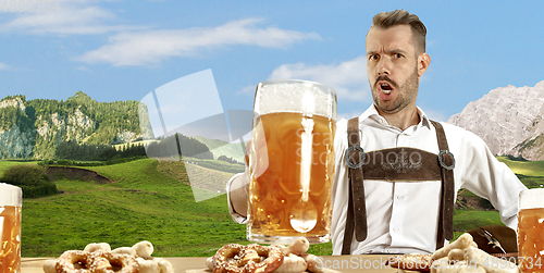 Image of The happy smiling man with beer dressed in traditional Austrian or Bavarian costume holding mug of beer, mountains on background, flyer