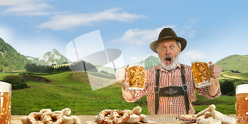 Image of The senior happy smiling man with beer dressed in traditional Austrian or Bavarian costume holding mug of beer, mountains on background, flyer