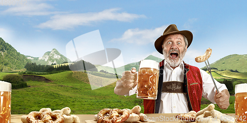 Image of The senior happy smiling man with beer dressed in traditional Austrian or Bavarian costume holding mug of beer, mountains on background, flyer