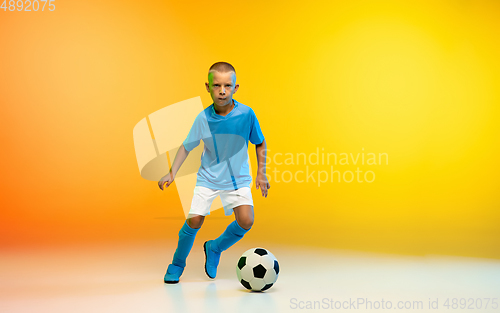 Image of Young boy as a soccer or football player in sportwear practicing on gradient yellow studio background in neon light