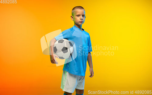Image of Young boy as a soccer or football player in sportwear practicing on gradient yellow studio background in neon light