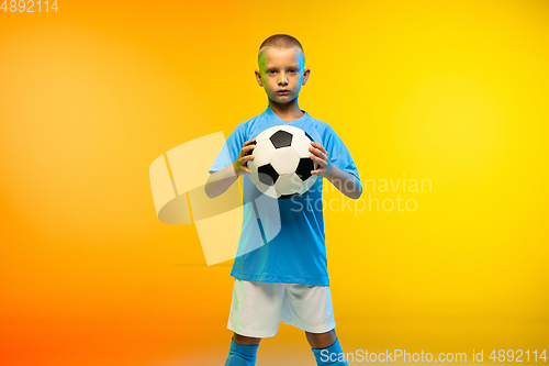 Image of Young boy as a soccer or football player in sportwear practicing on gradient yellow studio background in neon light