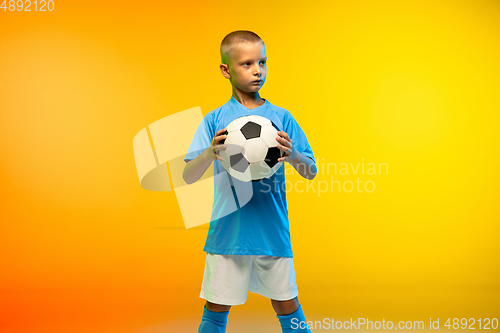 Image of Young boy as a soccer or football player in sportwear practicing on gradient yellow studio background in neon light